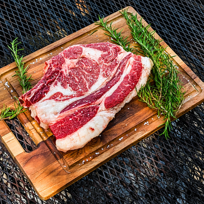 A raw marbled American Wagyu ribeye steak rests on a wooden cutting board with rosemary sprigs, placed on a round charcoal grill outdoors.