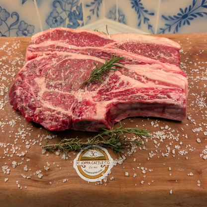 A raw American Wagyu marbled ribeye with visible bone, placed on a wooden cutting board sprinkled with coarse salt and rosemary, with a blue-and-white patterned tile wall behind it. A circular logo at the bottom reads 3C Joppa Cattle Co