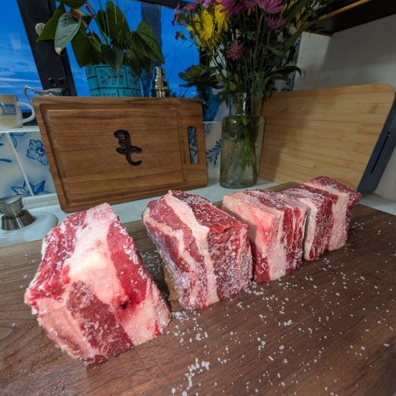 Four raw american wagyu short ribs sections with visible bone and fat marbling, arranged on a wooden cutting board in a kitchen setting with salt crystals scattered around. A wooden cutting board with a black bear emblem and flowers in the background are also visible.